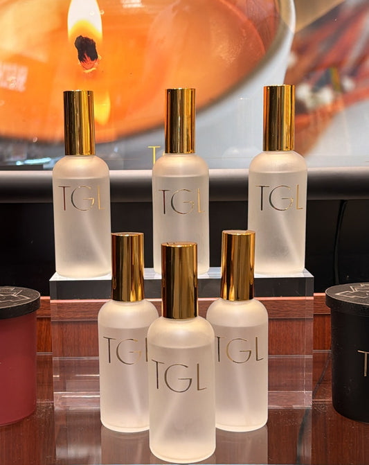 Five frosted glass bottles with gold caps and gold 'TGL' lettering, arranged on a reflective surface with a warm, blurred orange background.The GOOD LIFE Room Spray, linen spray