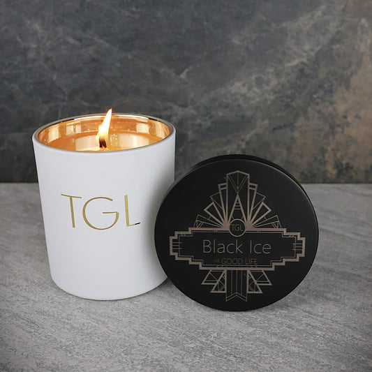 White candle with gold interior and lid labeled 'TGL' next to a black coaster with 'Black Ice' design on a gray surface.