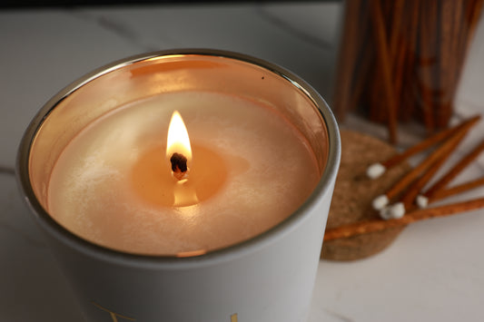 Soy Candle in a white container with a gold rim, lit with gold letters 'TGL' on a neutral background. Soy candle TGL - The GOOD LIFE
