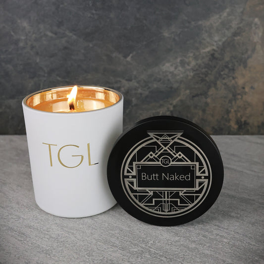 Candle with 'TGL' branding and a coaster with 'Butt Naked' text on a gray surface.