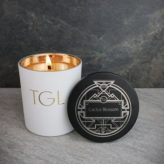 White candle with gold interior and black lid featuring 'TGL' and 'Cactus Blossom' on a gray surface.