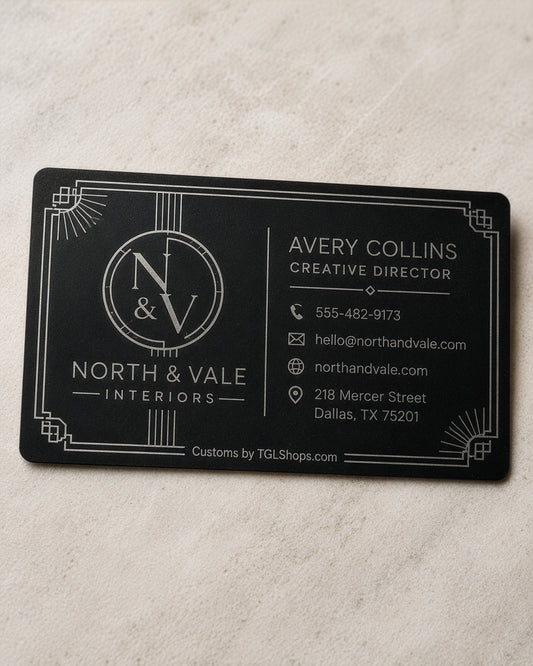 A rectangular black business card with white Art Deco-style border details and printed contact information for North & Vale Interiors, placed on a light beige textured surface.