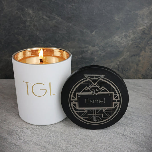 White candle with gold interior and black lid labeled 'TGL' on a gray surface with a dark background.