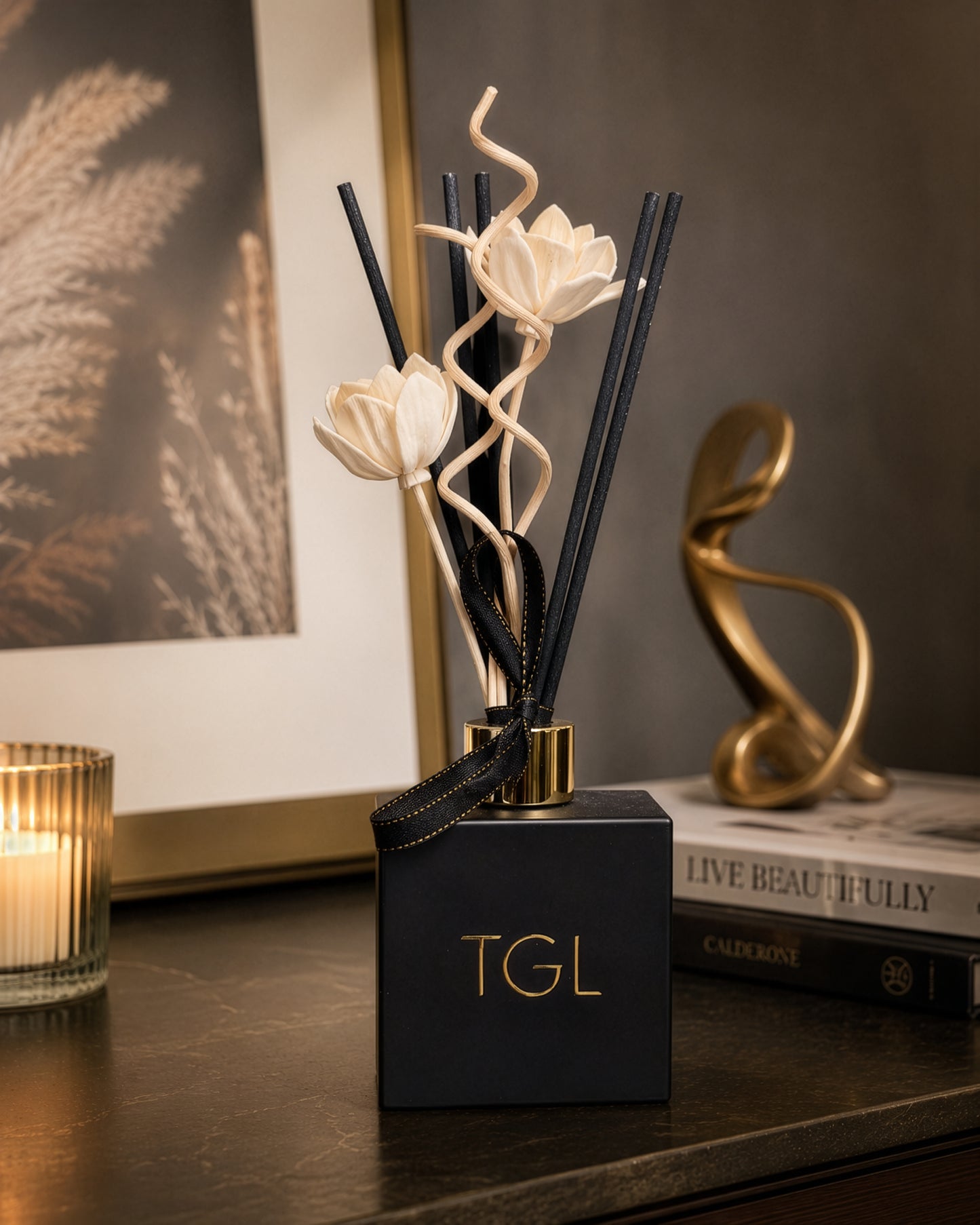 A reed diffuser with a matte black square base, gold lettering reading 'TGL', black reeds, and two cream-colored lotus flower decorations, styled on a dark wooden table next to a candle and books.