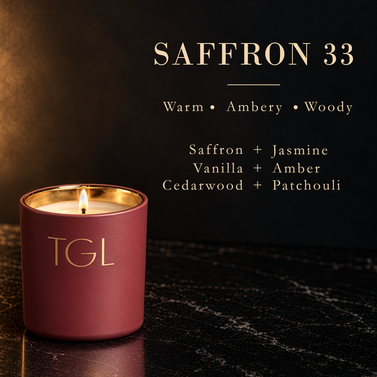 A lit TGL Saffron 33 scented candle in a matte burgundy jar with gold lettering and a gold inner rim, set on a dark marble surface against a dark background with product text.