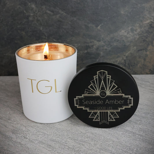White candle with gold interior and black lid labeled 'TGL' on a gray surface with dark background
