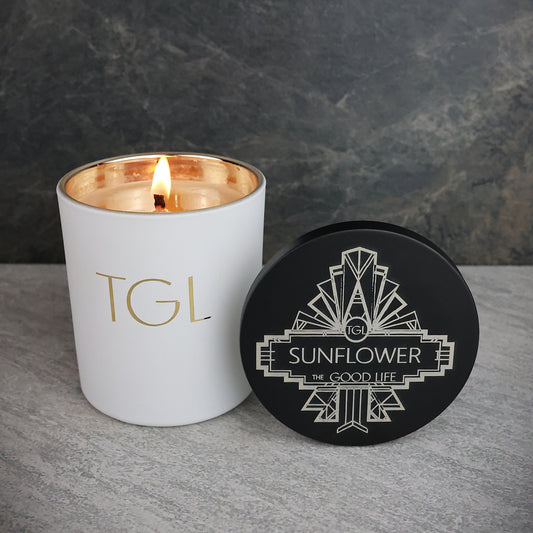 White candle with gold interior and black lid featuring 'TGL' branding on a gray surface with dark background