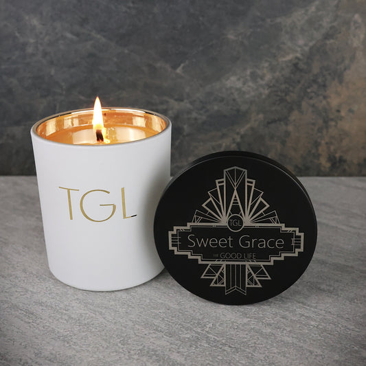 White candle with gold interior and 'TGL' branding next to a black coaster with 'Sweet Grace' branding on a gray surface.