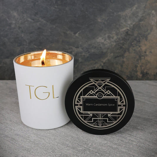 White candle with gold interior and 'TGL' branding next to a black coaster on a gray surface.