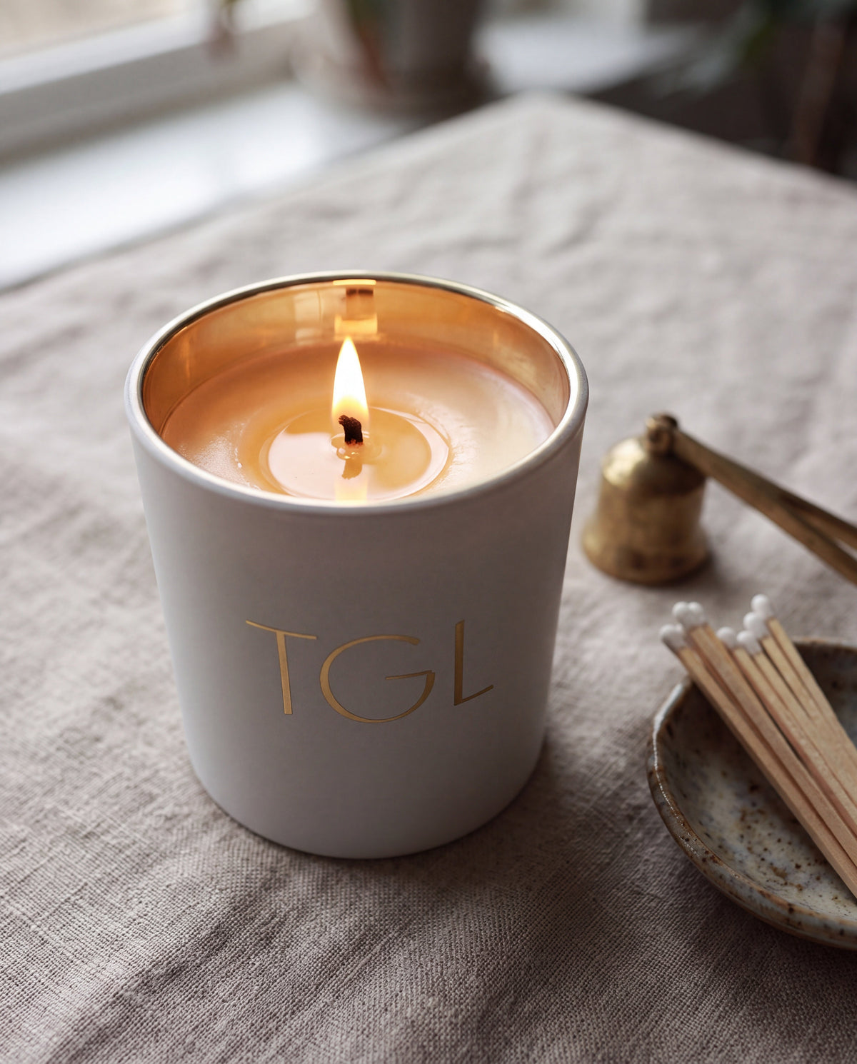 TGL soy candle by The GOOD LIFE, lit in a white jar on a linen table beside incense sticks