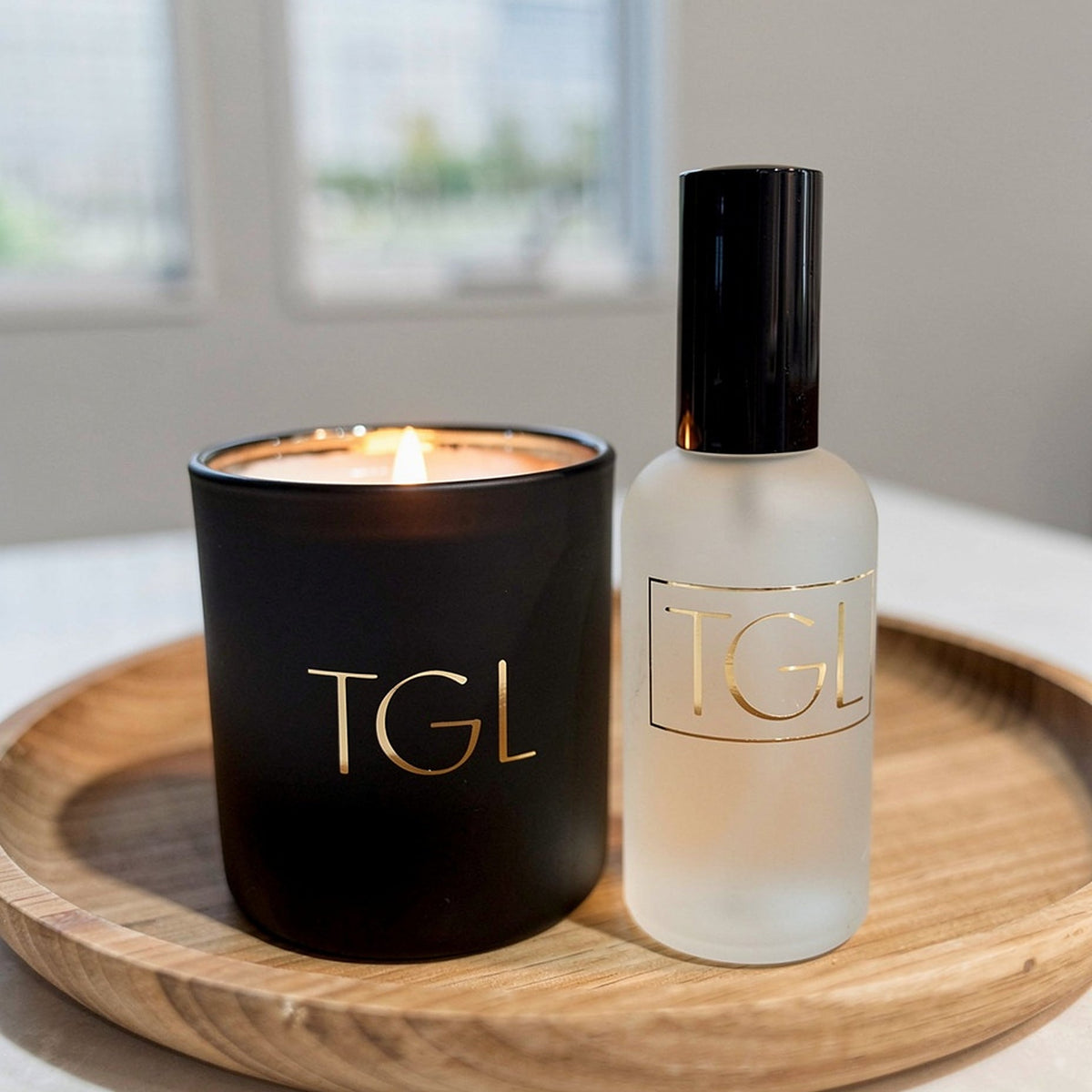 TGL soy candle by The GOOD LIFE, lit in a black jar beside a frosted room spray on a wooden tray