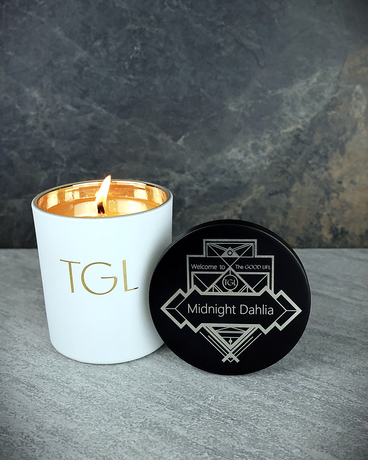 Lit Midnight Dahlia TGL soy candle beside its black lid engraved with an Art Deco design and the scent name