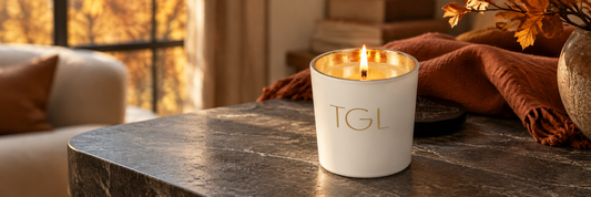 Lit white TGL candle on a wooden table with autumn leaves and a rust throw in warm fall light