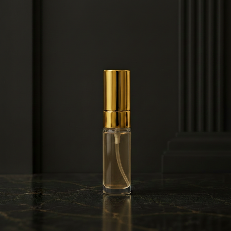 A small clear glass perfume atomizer bottle with a shiny gold metallic cap, standing on a dark marble surface against a beige wall with architectural molding.