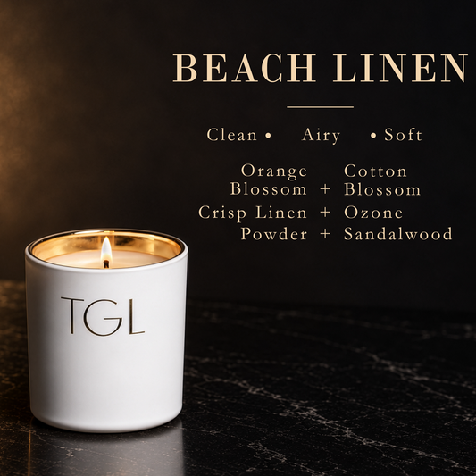 A lit white TGL candle labeled 'BEACH LINEN' sits on a dark marble surface against a dark background, with text describing the scent notes: Clean, Airy, Soft, Orange, Cotton, Blossom, Crisp Linen + Ozone, Powder, and Sandalwood.