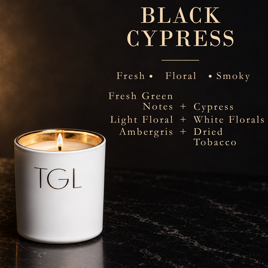 A white TGL candle with a gold-wrapped rim and lit wick sits on a dark marble surface against a black background. Gold text at the top reads 'BLACK CYPRESS' with scent notes listed below, including fresh green, floral, smoky, ambergris, and dried tobacco.