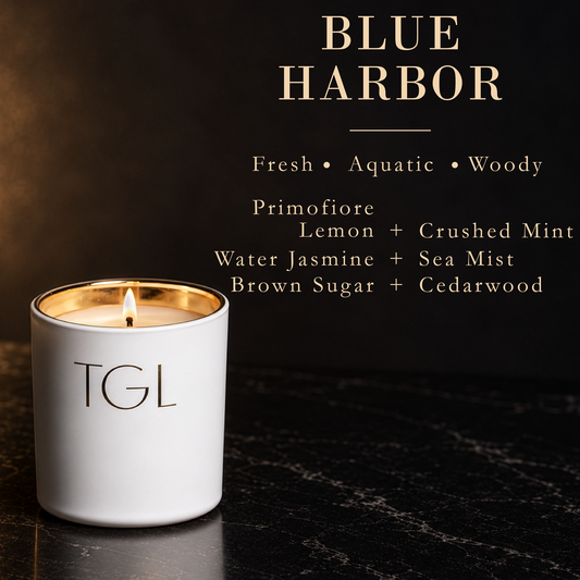 A white TGL candle with a gold-wrapped inner rim and a lit wick sits on a dark marble surface against a dark background. Gold text at the top reads 'BLUE HARBOR' with scent notes below, including Fresh, Aquatic, Woody, and ingredient mentions.