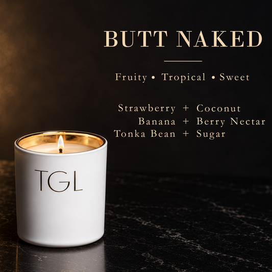A white TGL candle with a gold-wrapped rim and a lit wick sits on a dark marble surface against a dark background with text reading 'BUTT NAKED' and scent notes: Fruity • Tropical • Sweet, Strawberry + Coconut, Banana + Berry Nectar, Tonka Bean + Sugar.