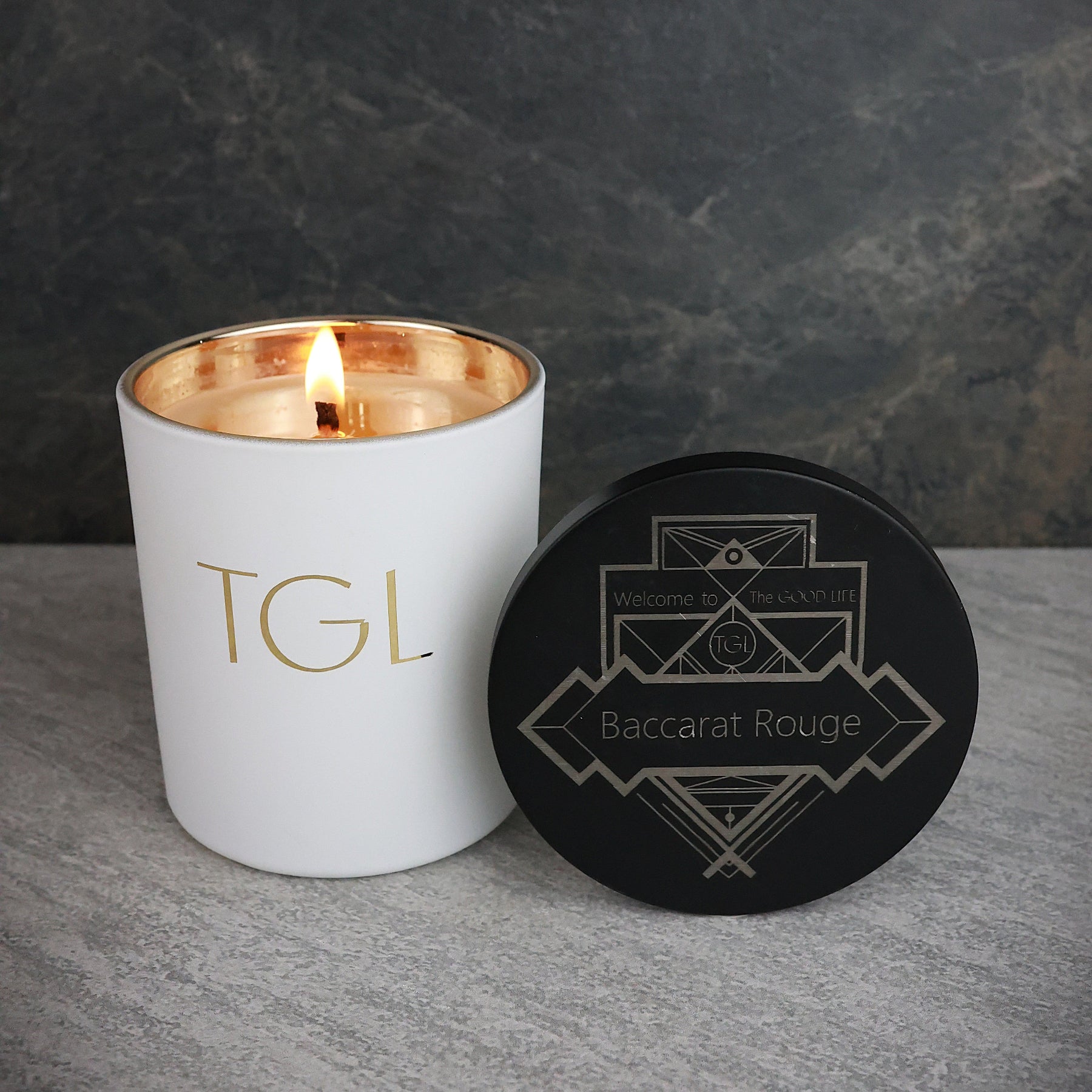 White candle with gold interior and black lid featuring geometric design on a gray surface. Baccarat Rouge candle natural soy beeswax blend 8oz