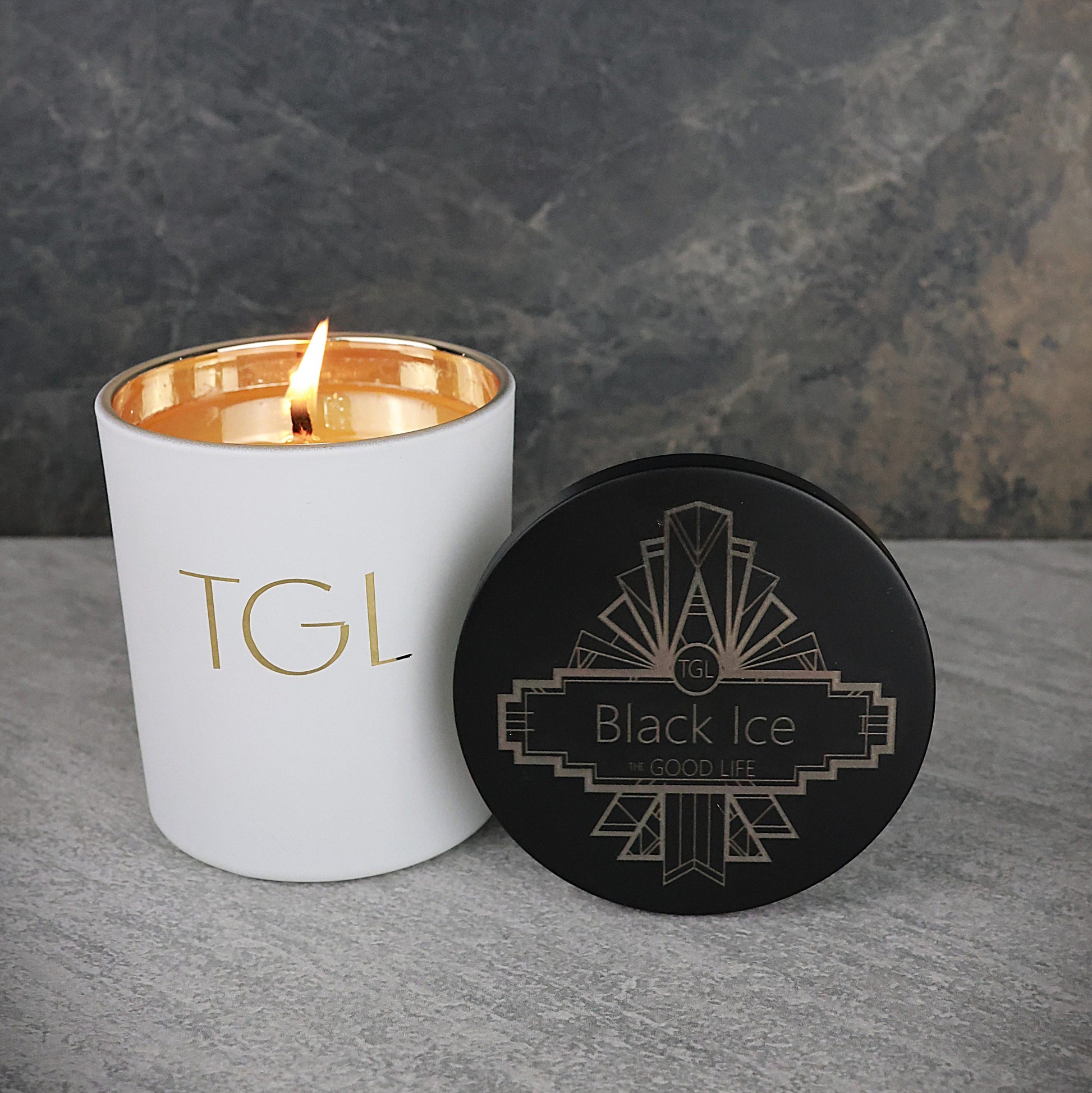 White candle with gold interior and lid labeled 'TGL' next to a black coaster with 'Black Ice' design on a gray surface.