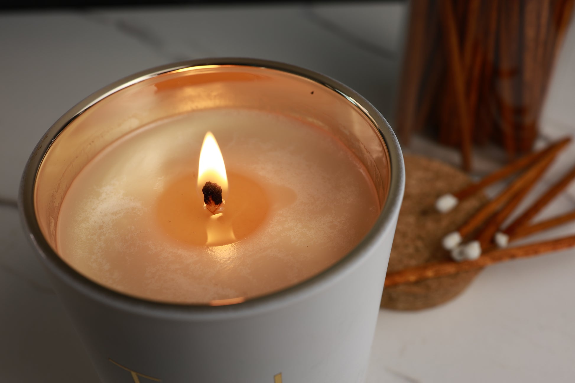 Soy Candle in a white container with a gold rim, lit with gold letters 'TGL' on a neutral background. Soy candle TGL - The GOOD LIFE