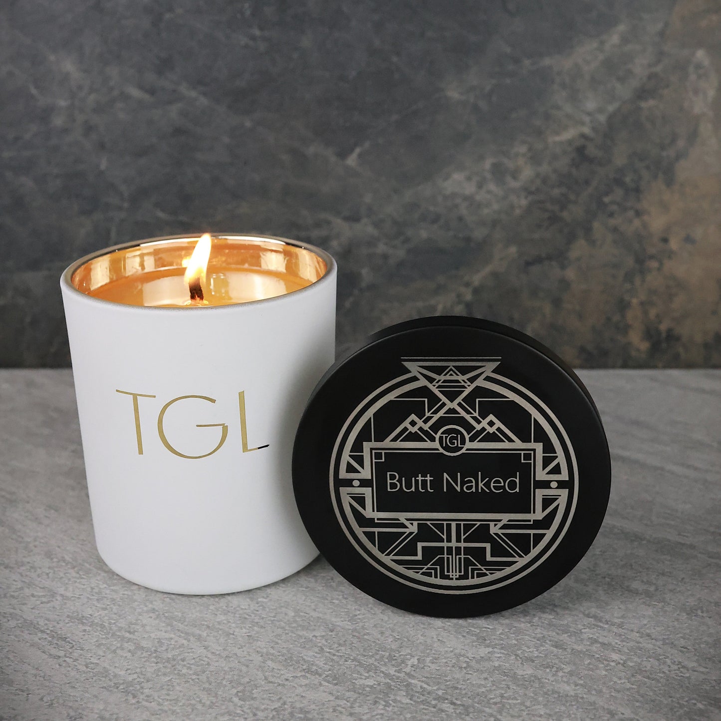 Candle with 'TGL' branding and a coaster with 'Butt Naked' text on a gray surface.