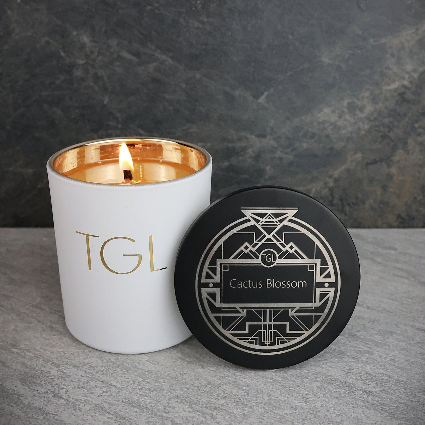 White candle with gold interior and black lid featuring 'TGL' and 'Cactus Blossom' on a gray surface.