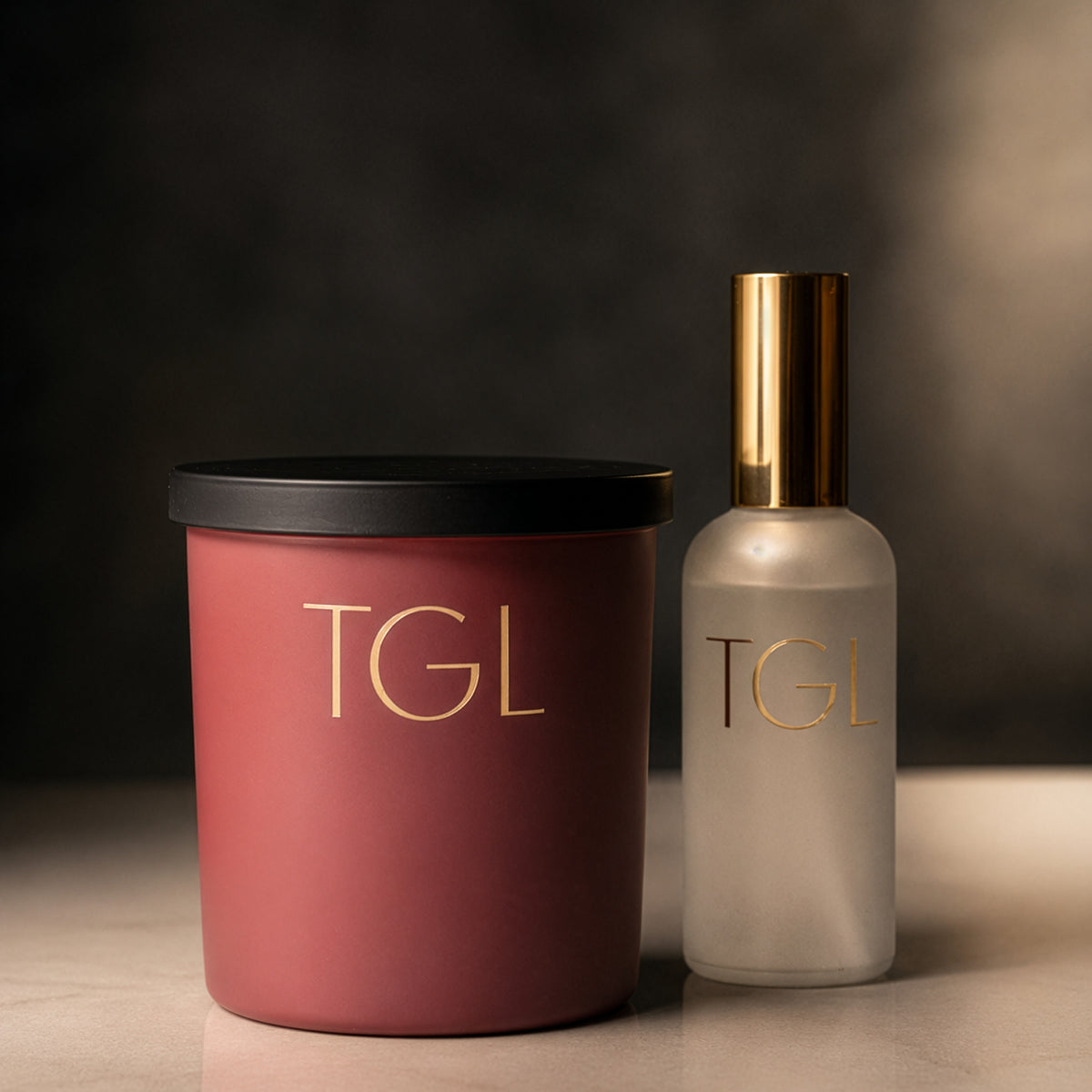 A matte rose soy saffron 33 candle with a black lid and gold 'TGL' lettering, next to a frosted glass saffron 33 room spray with a gold cap and gold 'TGL' lettering, on a light marble surface against a dark background.