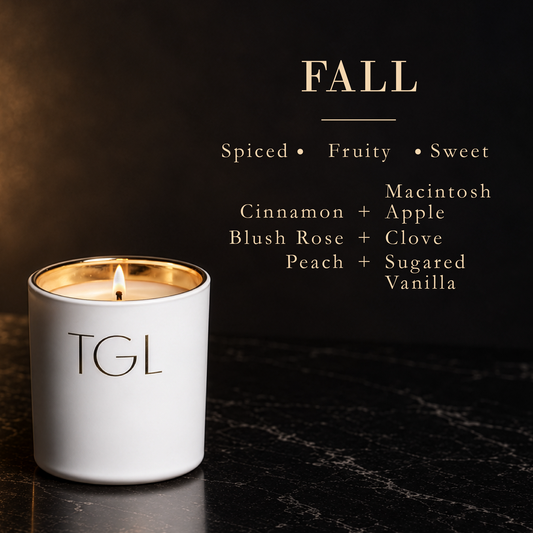 A white TGL candle with a gold inner rim and a lit wick sits on a dark marble surface against a dark background. Text on the image reads 'FALL' with scent descriptors 'Spiced • Fruity • Sweet' and lists notes including cinnamon, apple, blush rose, clove, peach, and sugared vanilla.
