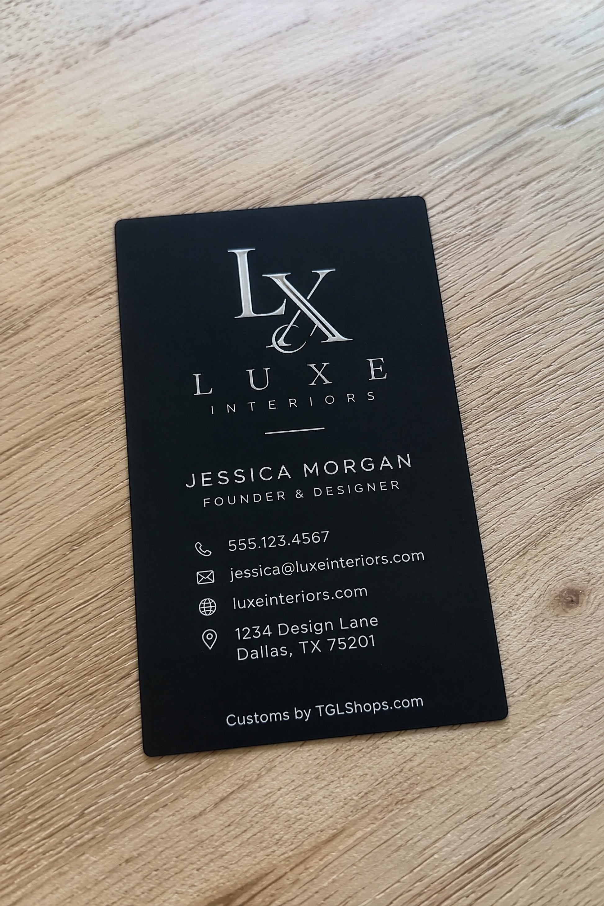 A black business card for Luxe Interiors by Jessica Morgan, placed on a light wood grain surface. The card features white text and logo details.