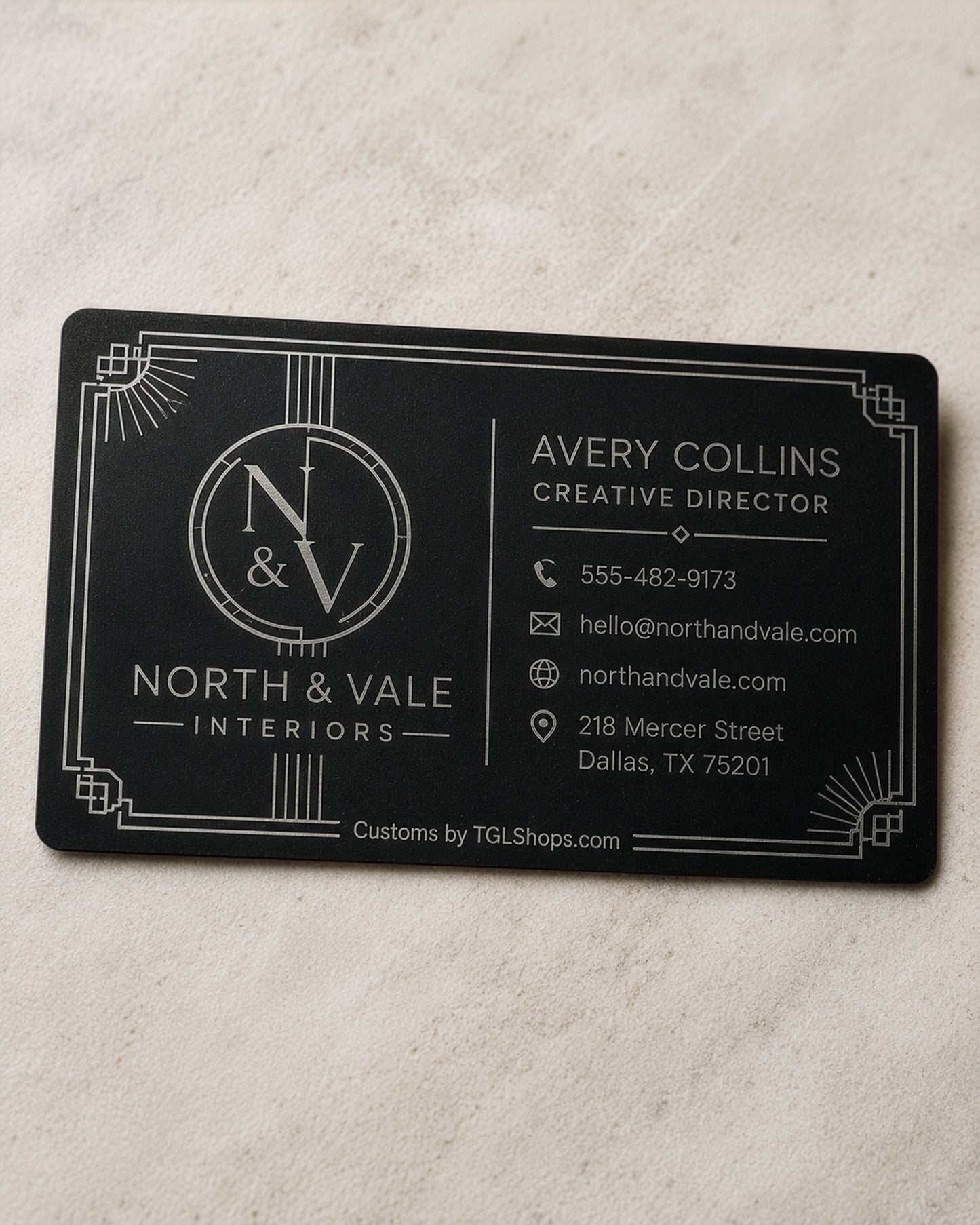 A rectangular black business card with white Art Deco-style border details and printed contact information for North & Vale Interiors, placed on a light beige textured surface.