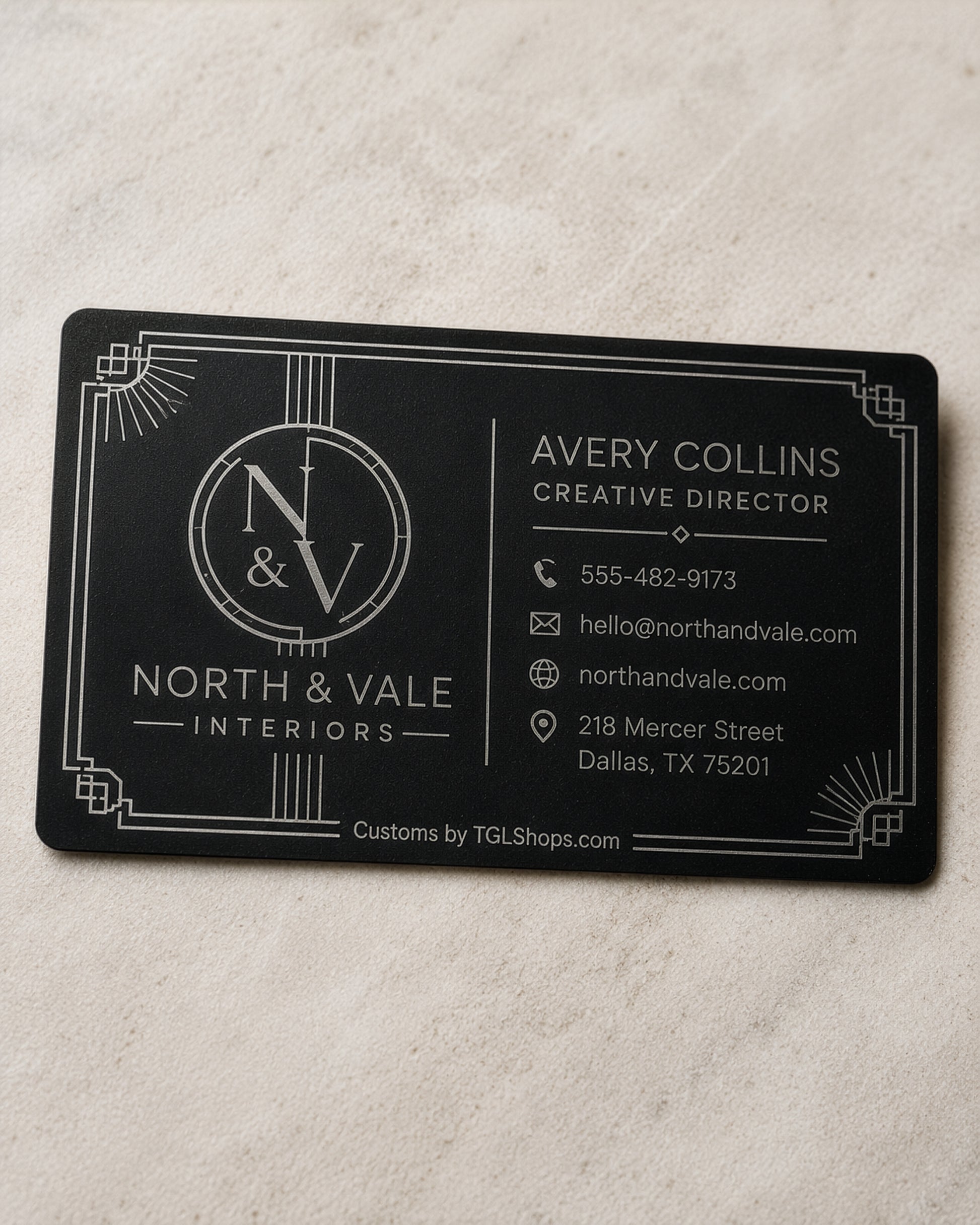 A rectangular black business card with white Art Deco-style border details and printed contact information for North & Vale Interiors, placed on a light beige textured surface.