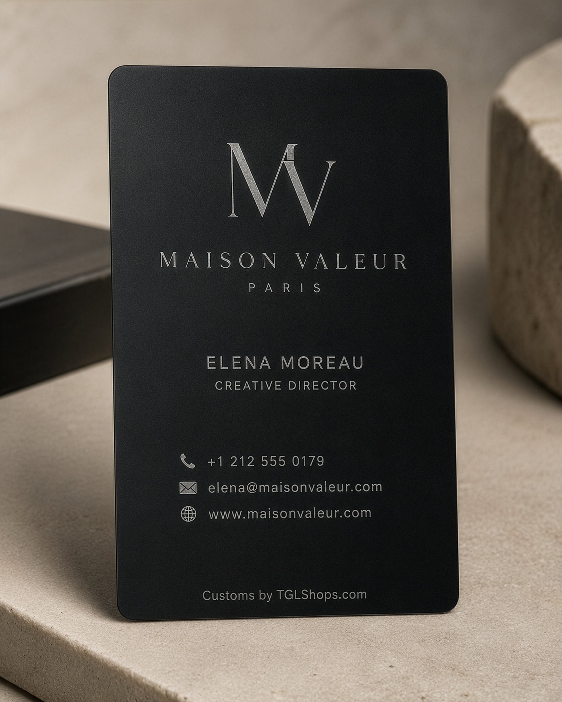 A matte black business card for Maison Valeur Paris, featuring elegant silver typography and contact details, displayed on a neutral beige stone surface.