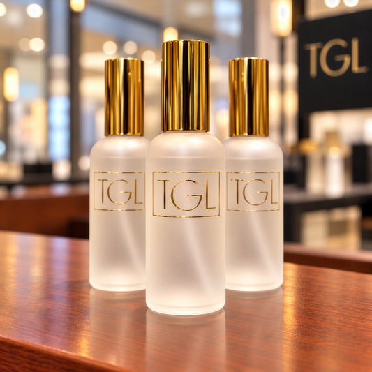 Three frosted glass skincare bottles with shiny gold caps and gold 'TGL' lettering, arranged on a polished wooden counter in a softly blurred spa or retail interior.
