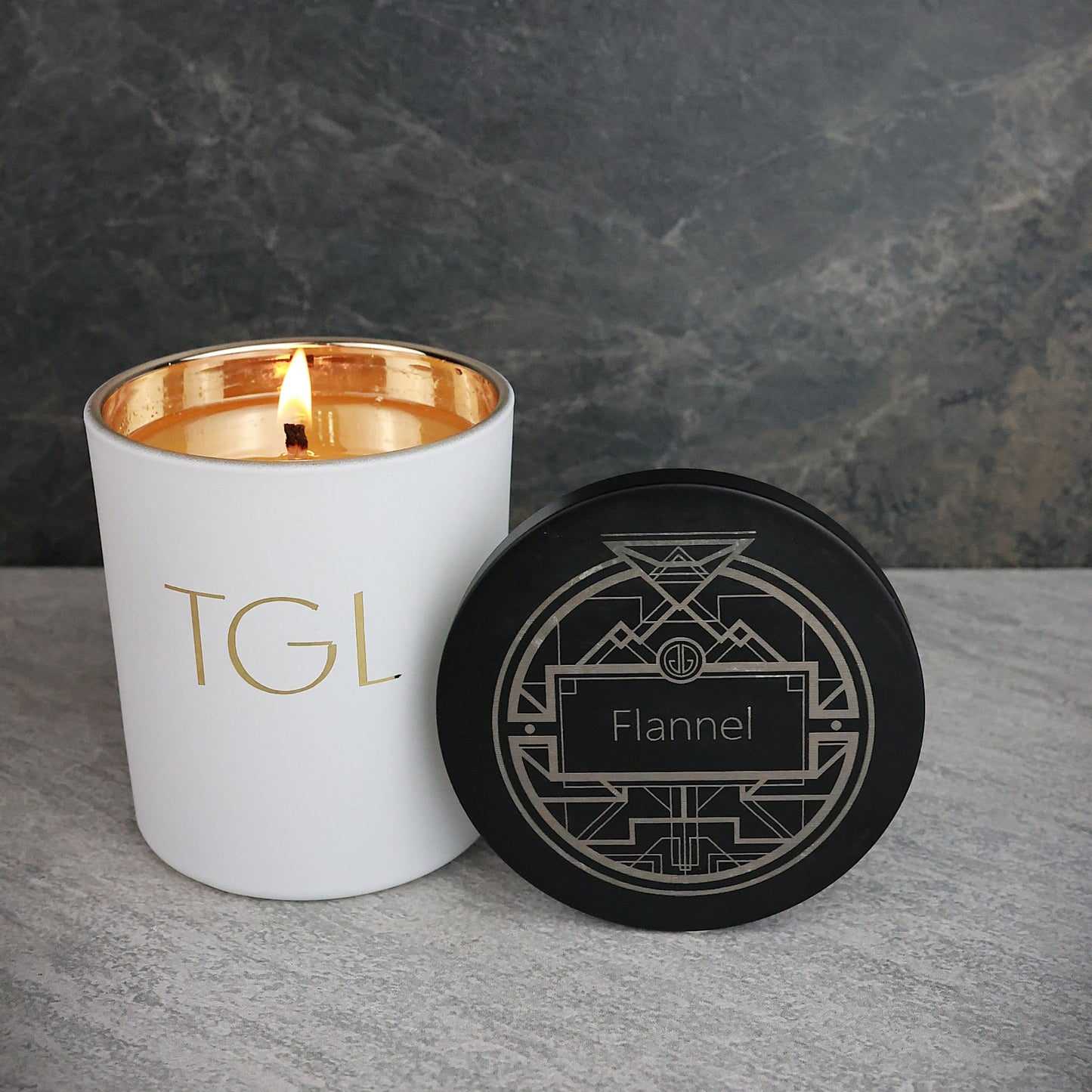 White candle with gold interior and black lid labeled 'TGL' on a gray surface with a dark background.