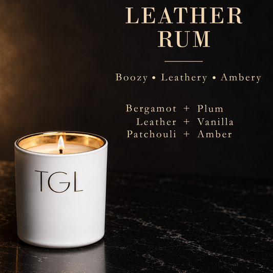 A white TGL candle with a gold rim and lit wick sits on a dark marble surface against a dark background. Text at the top reads 'LEATHER RUM' with scent notes 'Boozy • Leathery • Ambery' and ingredient descriptions 'Bergamot + Plum', 'Leather + Vanilla', and 'Patchouli + Amber'.