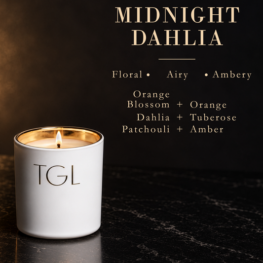 A white TGL candle with a gold-wrapped rim and a lit flame, set on a dark marble surface against a dark background with text reading 'MIDNIGHT DAHLIA' and scent notes.