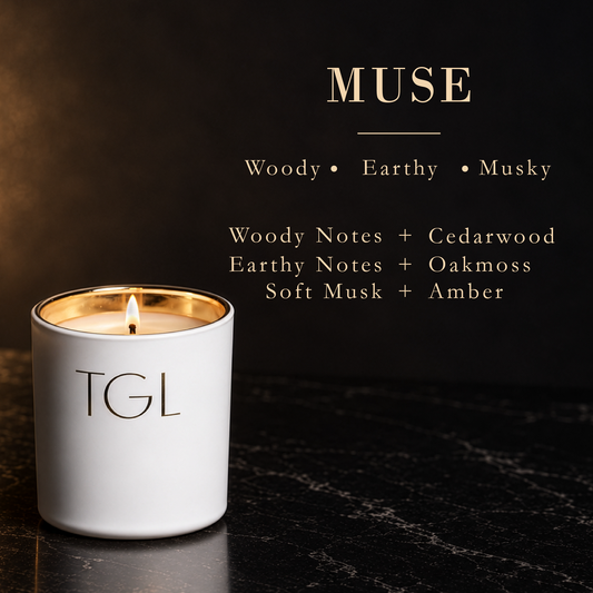 A white TGL candle with a gold inner rim and a lit wick sits on a dark marble surface against a dark background. Gold text above reads 'MUSE' with scent notes: Woody, Earthy, Musky, Woody Notes + Cedarwood, Earthy Notes + Oakmoss, Soft Musk + Amber.