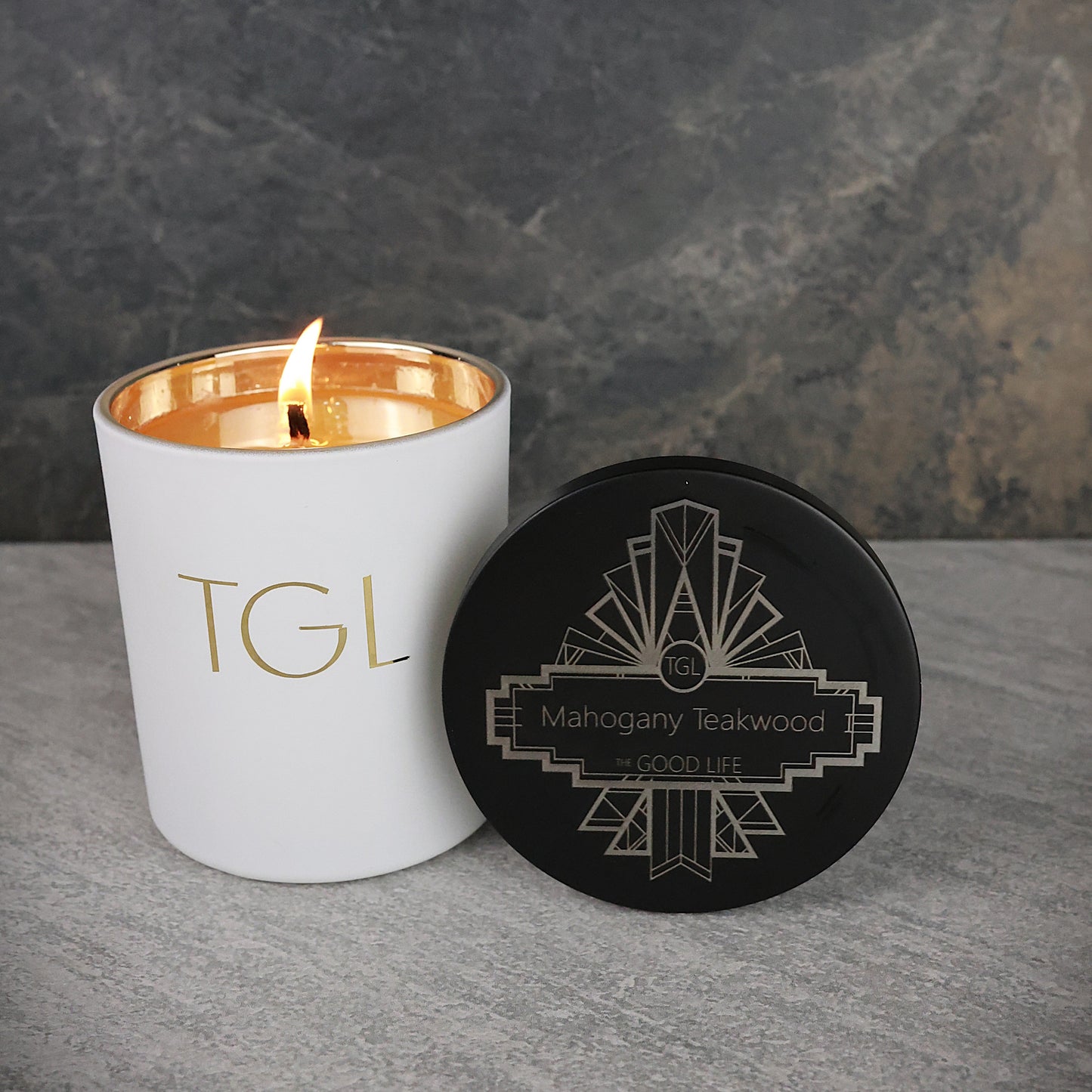 White candle with gold interior and black lid featuring 'TGL' branding on a gray surface.