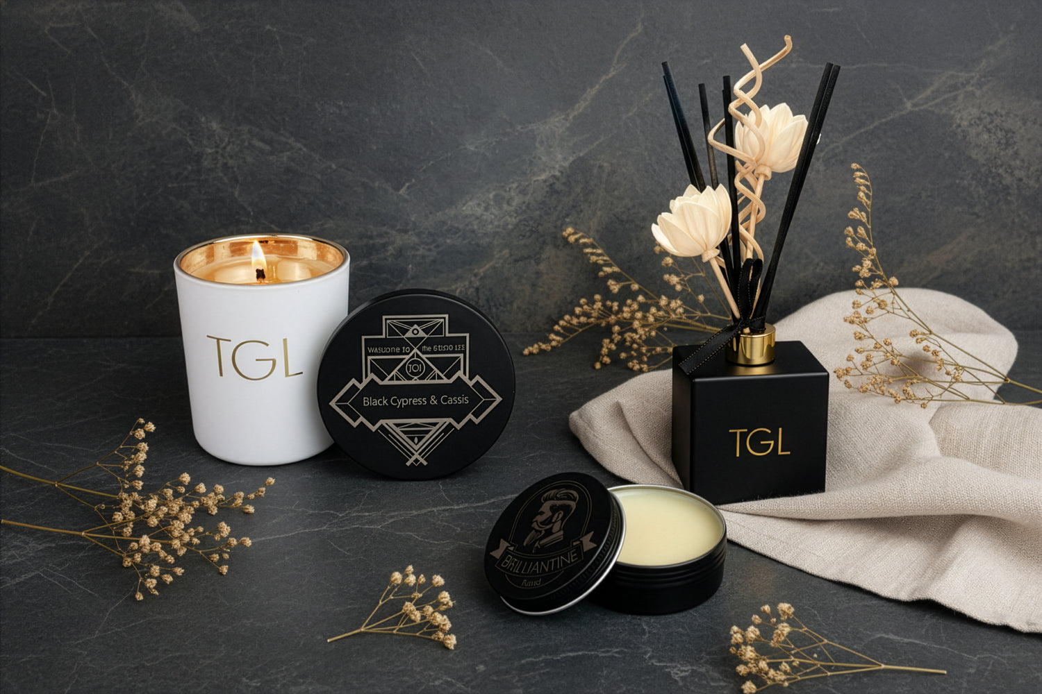 A styled product arrangement on a dark stone surface featuring a white so candle from The GOOD LIFE. labeled TGL, The GOOD LIFE a black round tin with a The GOOD LIFE Brillantine beard balm, an open black tin with cream-colored beeswax, and a black reed diffuser with cream flowers bamboos reeds and a TGL The GOOG LIFE logo. Fragrance oil, diffuser oil, Dried botanical sprigs and a cream fabric are arranged around the products.