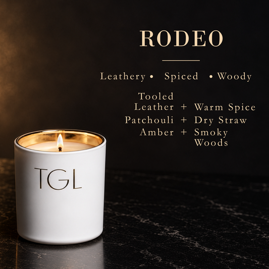 A lit white TGL candle with a gold rim sits on a dark marble surface against a dark background, with the word 'RODEO' and scent notes displayed above in gold text.