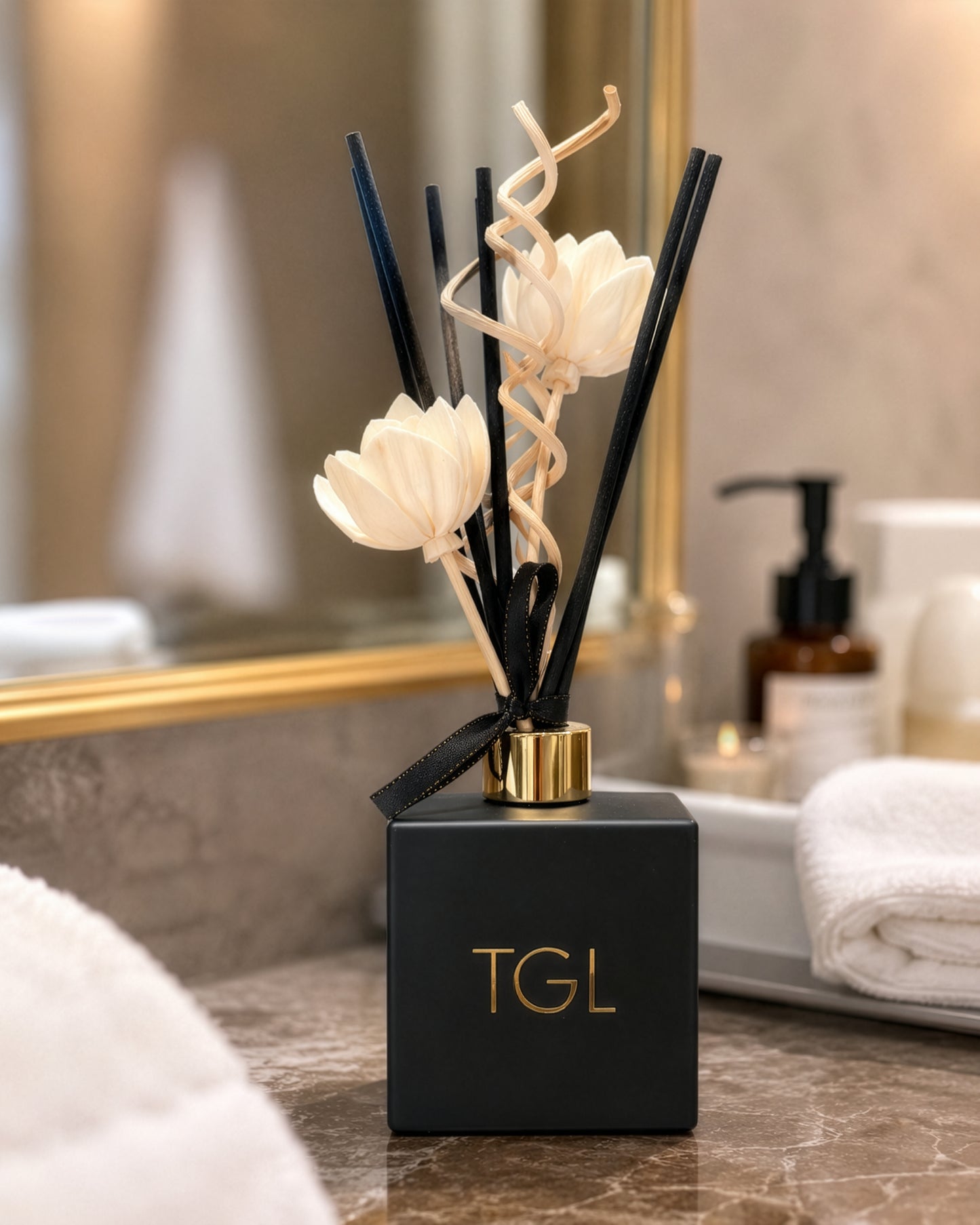 A luxury reed diffuser with a matte black square bottle, gold-embossed 'TGL' logo, and natural reeds decorated with white flower ornaments, set on a marble bathroom counter.