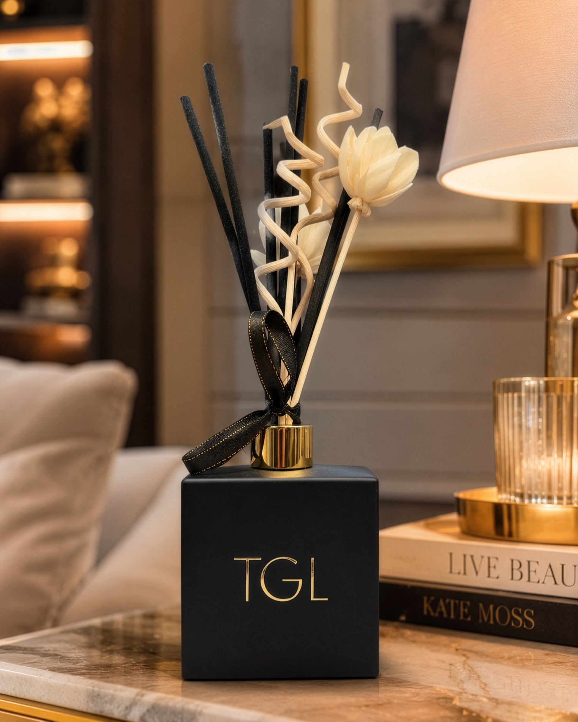 A luxury reed diffuser with a matte black square base, gold lettering, and natural reeds including twisted cream and black elements, styled on a marble table next to a lamp.