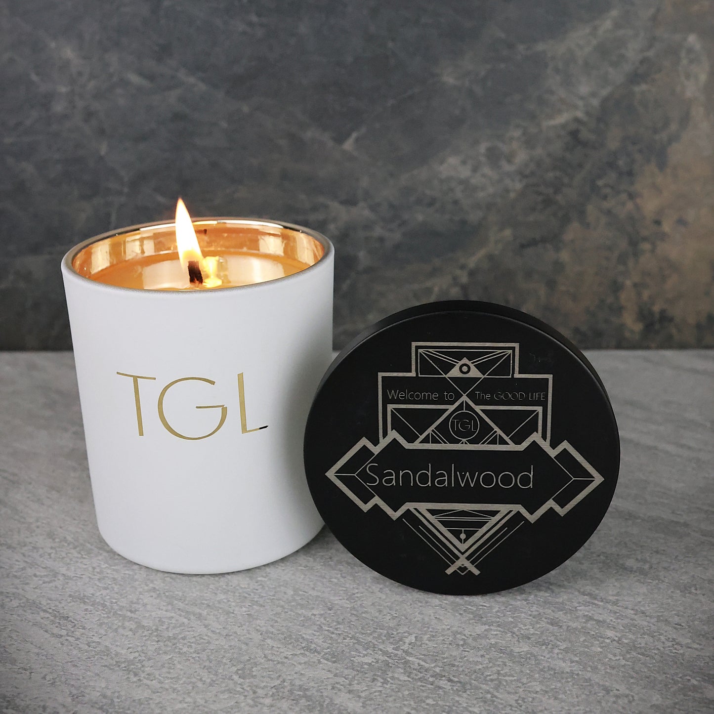 Candle with 'TGL' branding and a black coaster with 'Sandalwood' text on a gray surface.