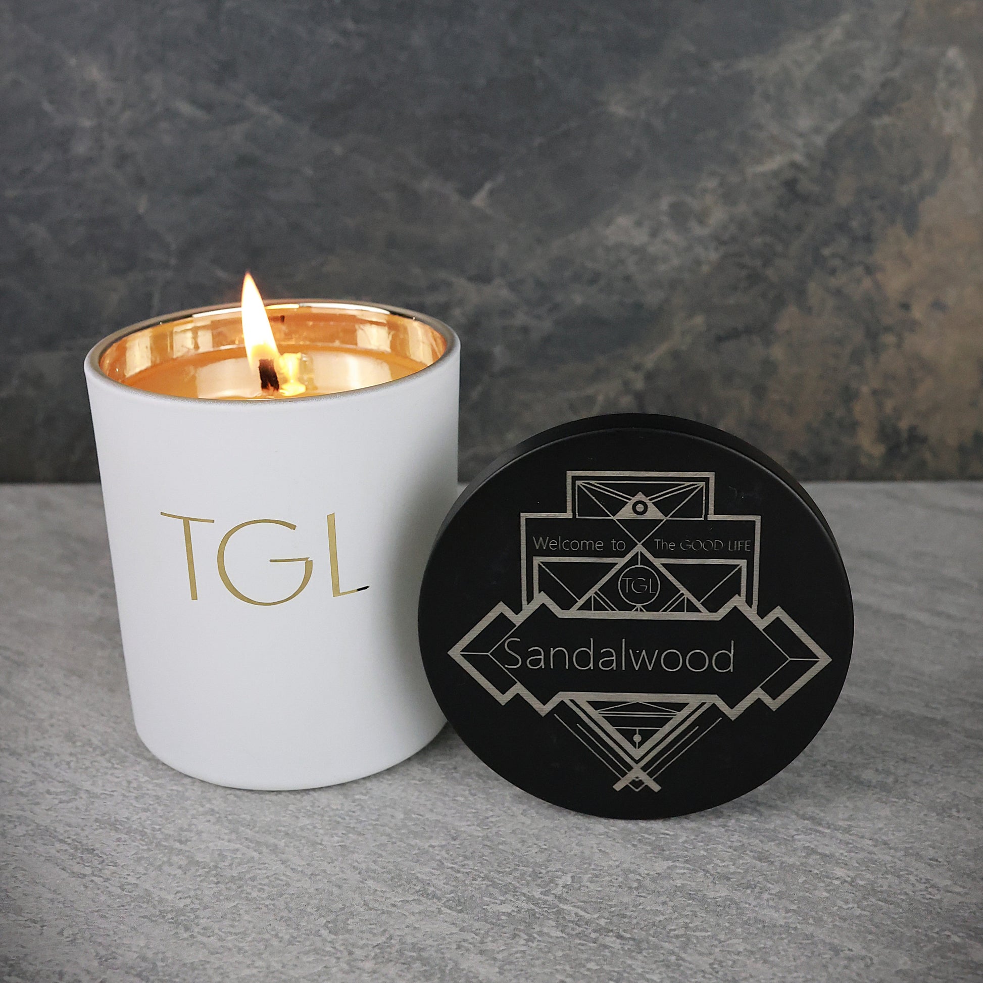 Candle with 'TGL' branding and a black coaster with 'Sandalwood' text on a gray surface.