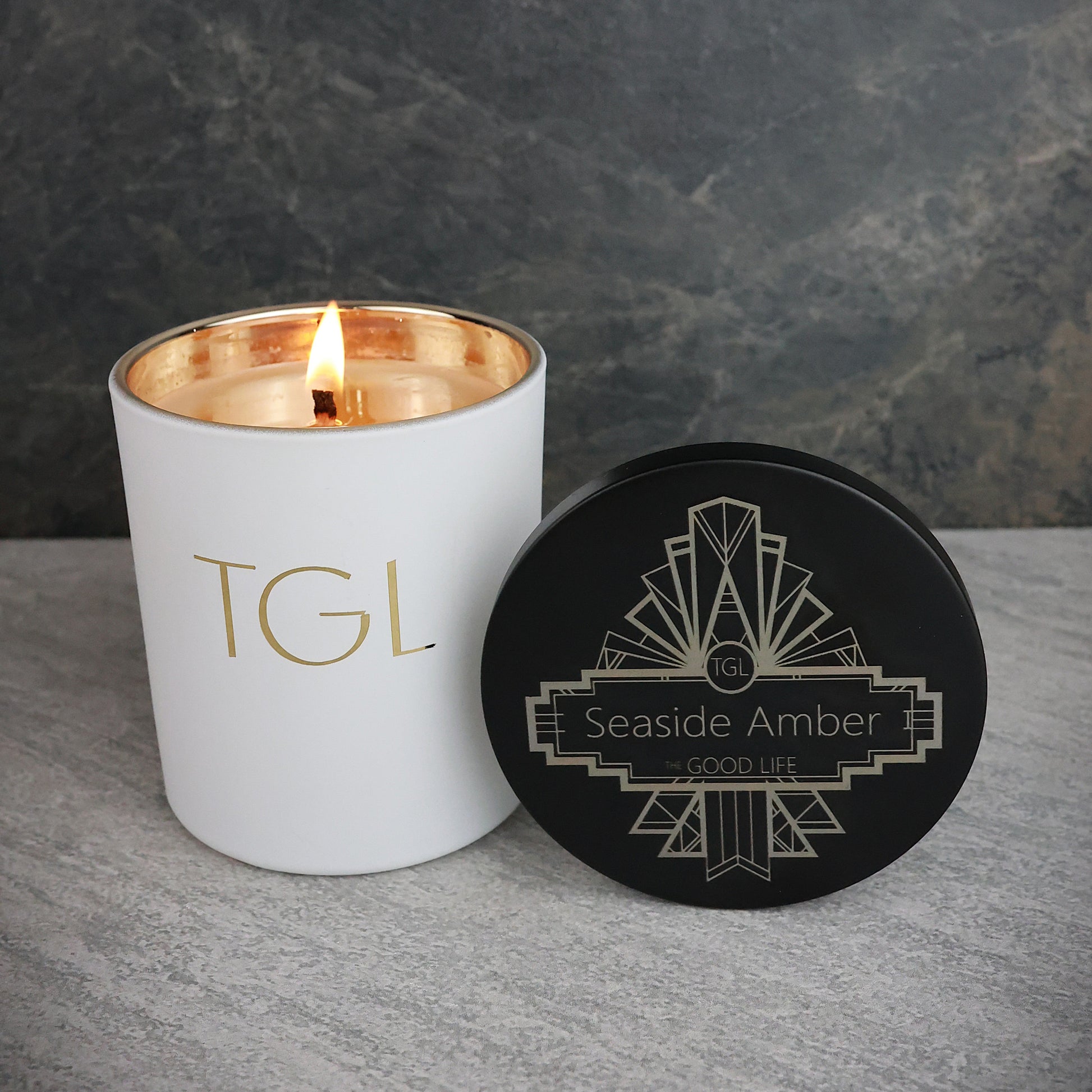 White candle with gold interior and black lid labeled 'TGL' on a gray surface with dark background