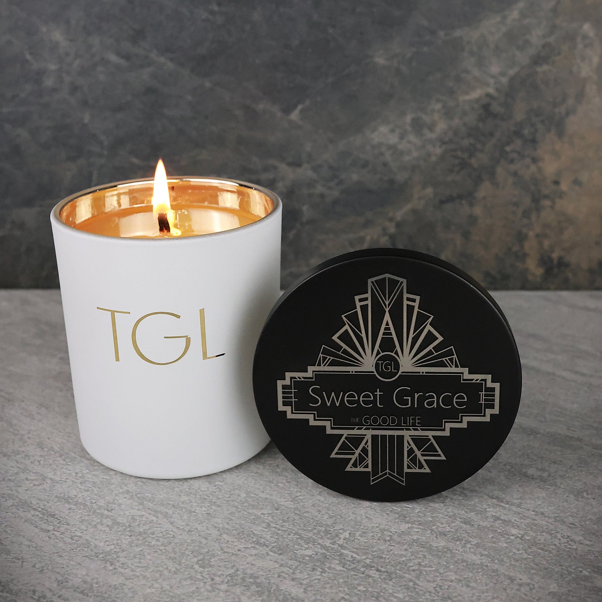 White candle with gold interior and 'TGL' branding next to a black coaster with 'Sweet Grace' branding on a gray surface.