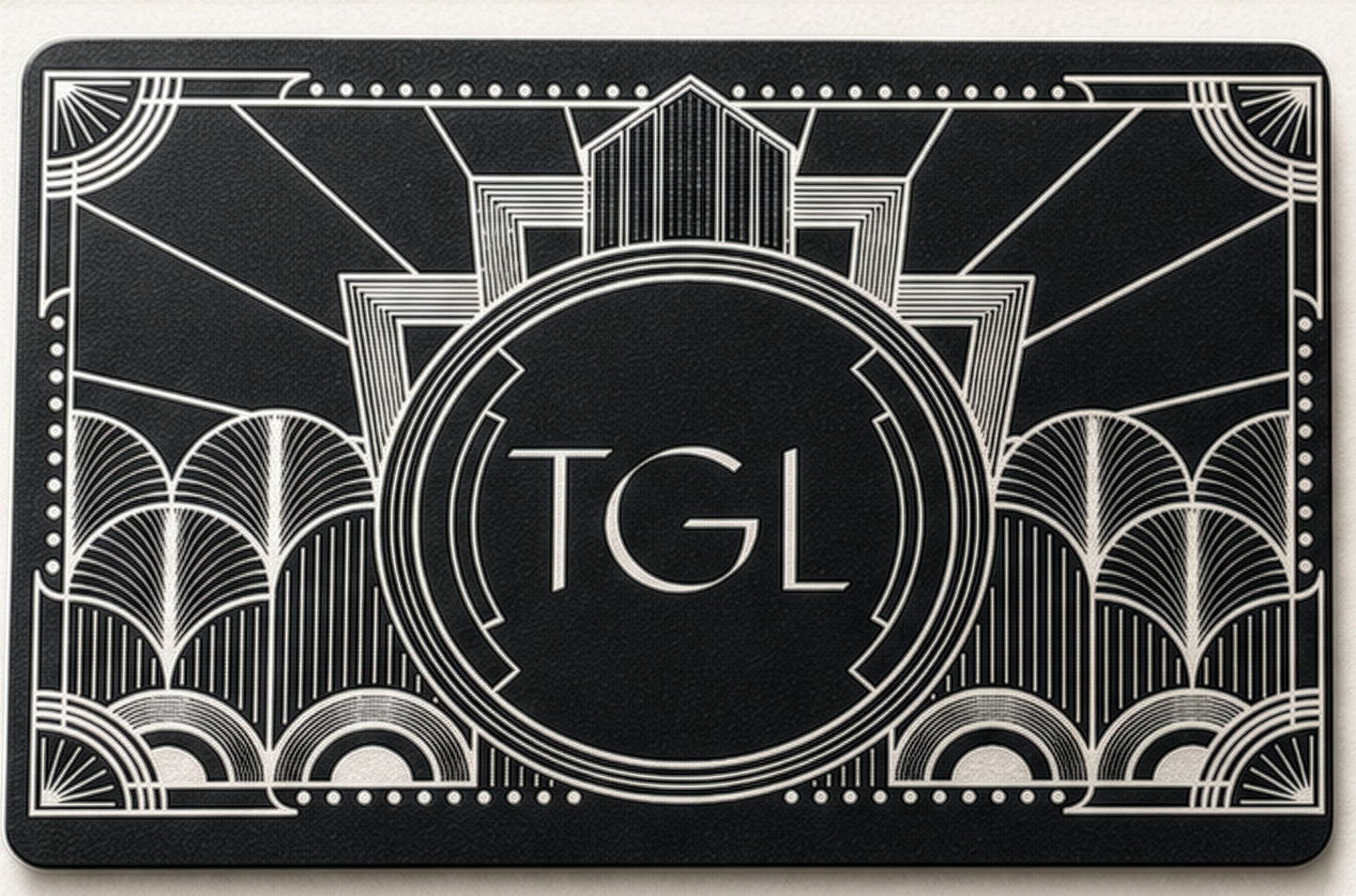 A black rectangular card or mat featuring an Art Deco style white line design with the letters 'TGL' centered inside a circular emblem.