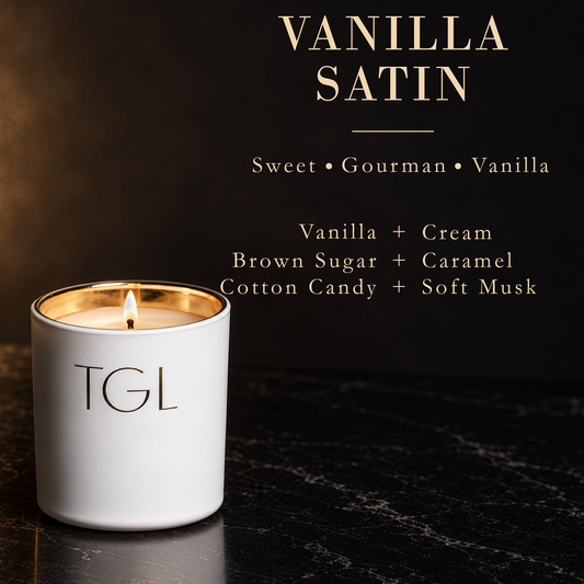 A white ceramic candle jar with a gold-toned inner rim and a lit wick, labeled 'TGL' on the front. The candle is set on a dark marble surface against a dark background with elegant gold text reading 'VANILLA SATIN' and scent notes including vanilla, cream, brown sugar, caramel, cotton candy, and soft musk.