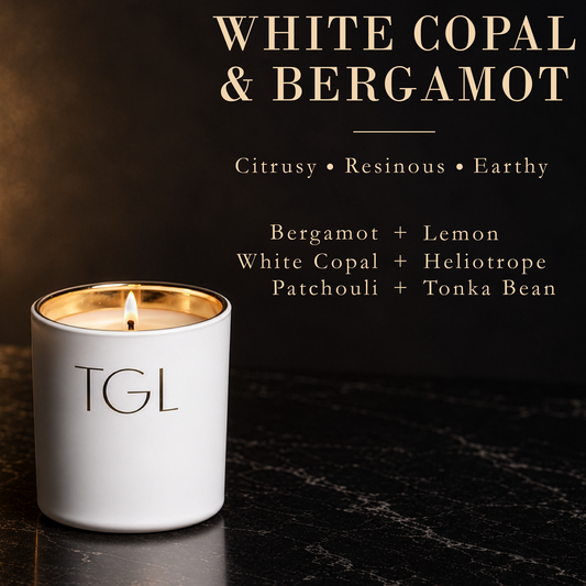 A white ceramic candle jar with a gold-toned inner rim and a lit flame, labeled TGL, on a dark marble surface. The image includes text reading 'WHITE COPAL & BERGAMOT' and scent notes: Citrusy • Resinous • Earthy, Bergamot + Lemon, White Copal + Heliotrope, Patchouli + Tonka Bean.